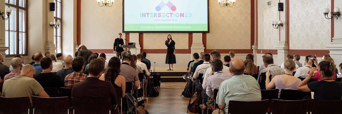 A speaker on stage in a grand hall, the Intersection 23 title slide behind her.
